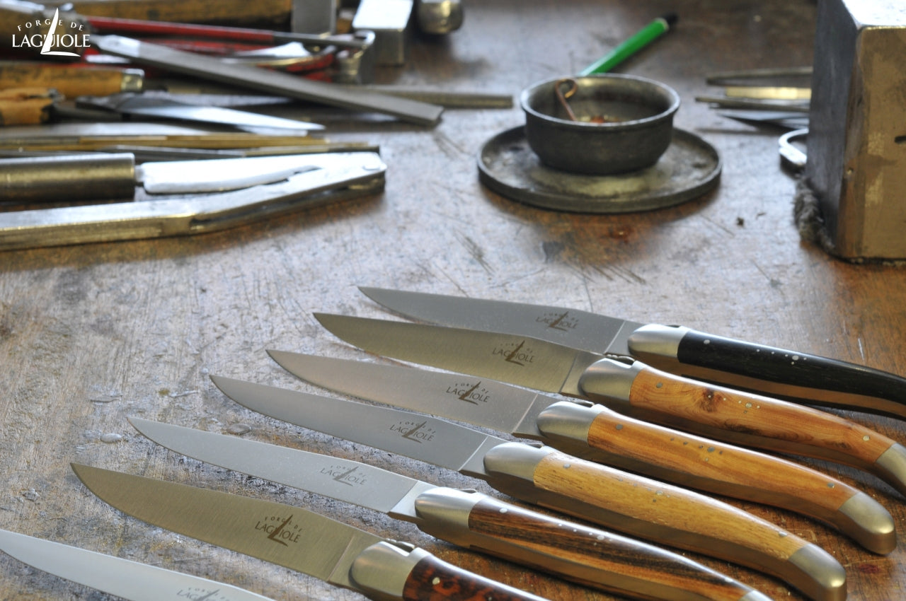 several forge de laguiole steak knives with mixed handle material on a wooden work bench