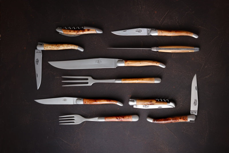 several cutlery items from forge de laguiole on a black background. table knife, pocket knife, carving knife and fork and a waiter style corkscrew on the top left.