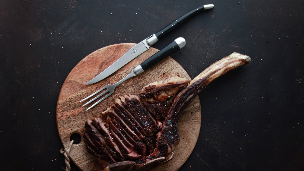 Forge de Laguiole ebony handle carving set on a round cutting board next to a Tomahawk steak