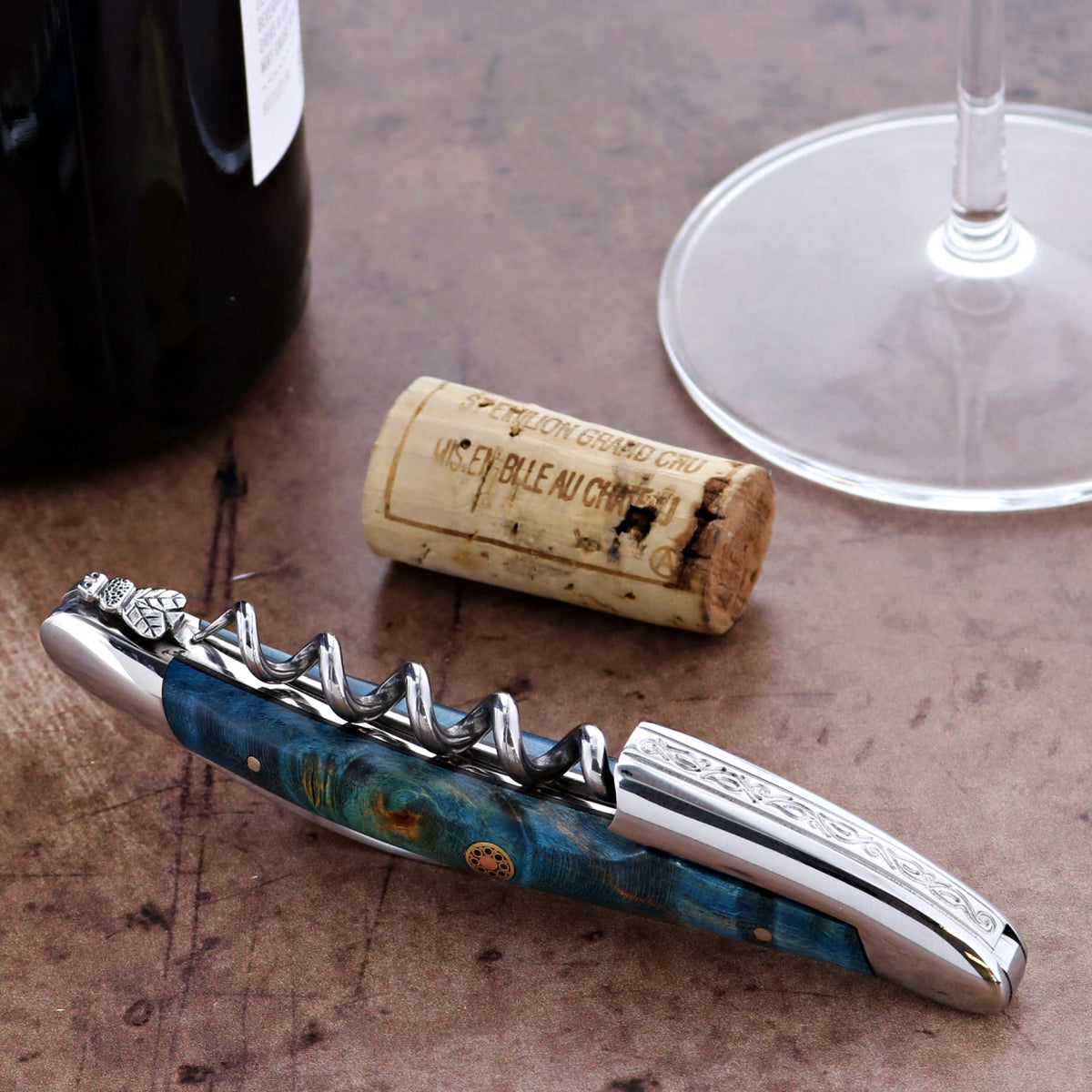 Wine opener with blue handle and silver accents on a wooden surface with a cork and glass.