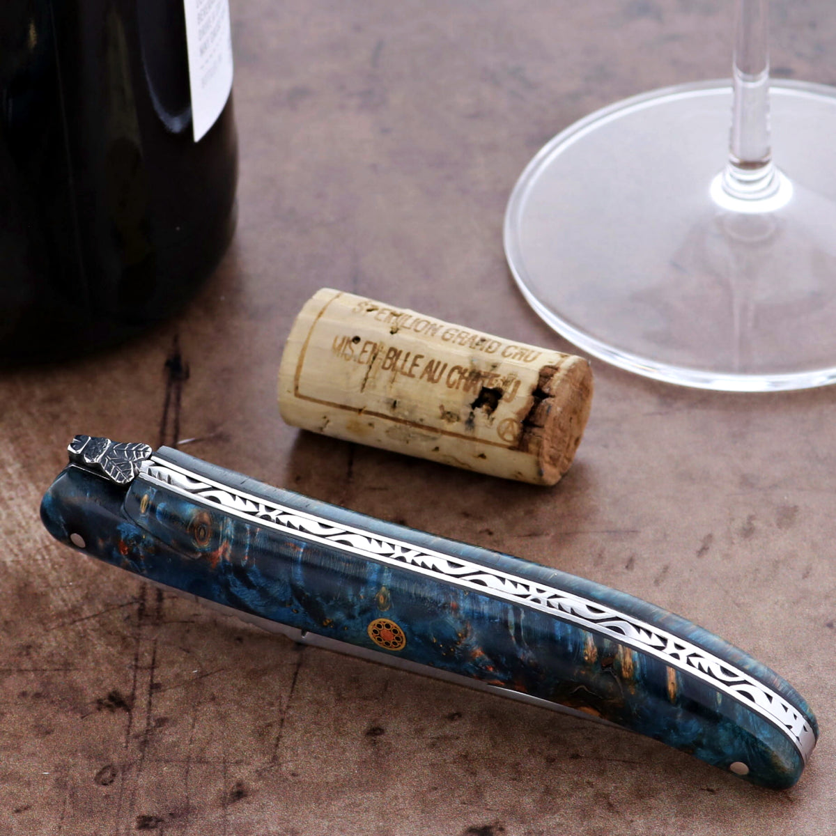 Blue folding knife on a wooden surface with a cork and glass in the background