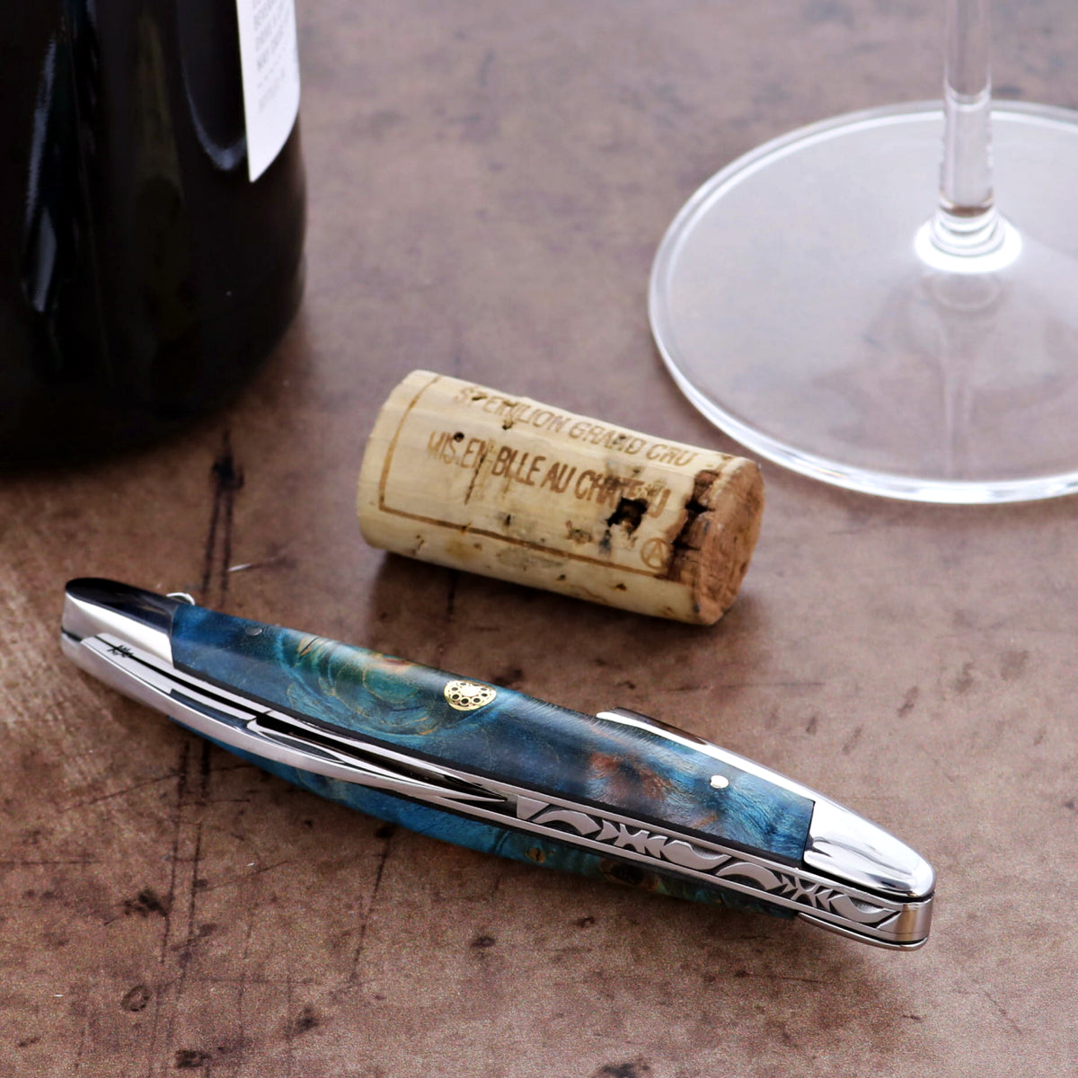 Blue corkscrew on a wooden surface with a wine cork and glass in the background