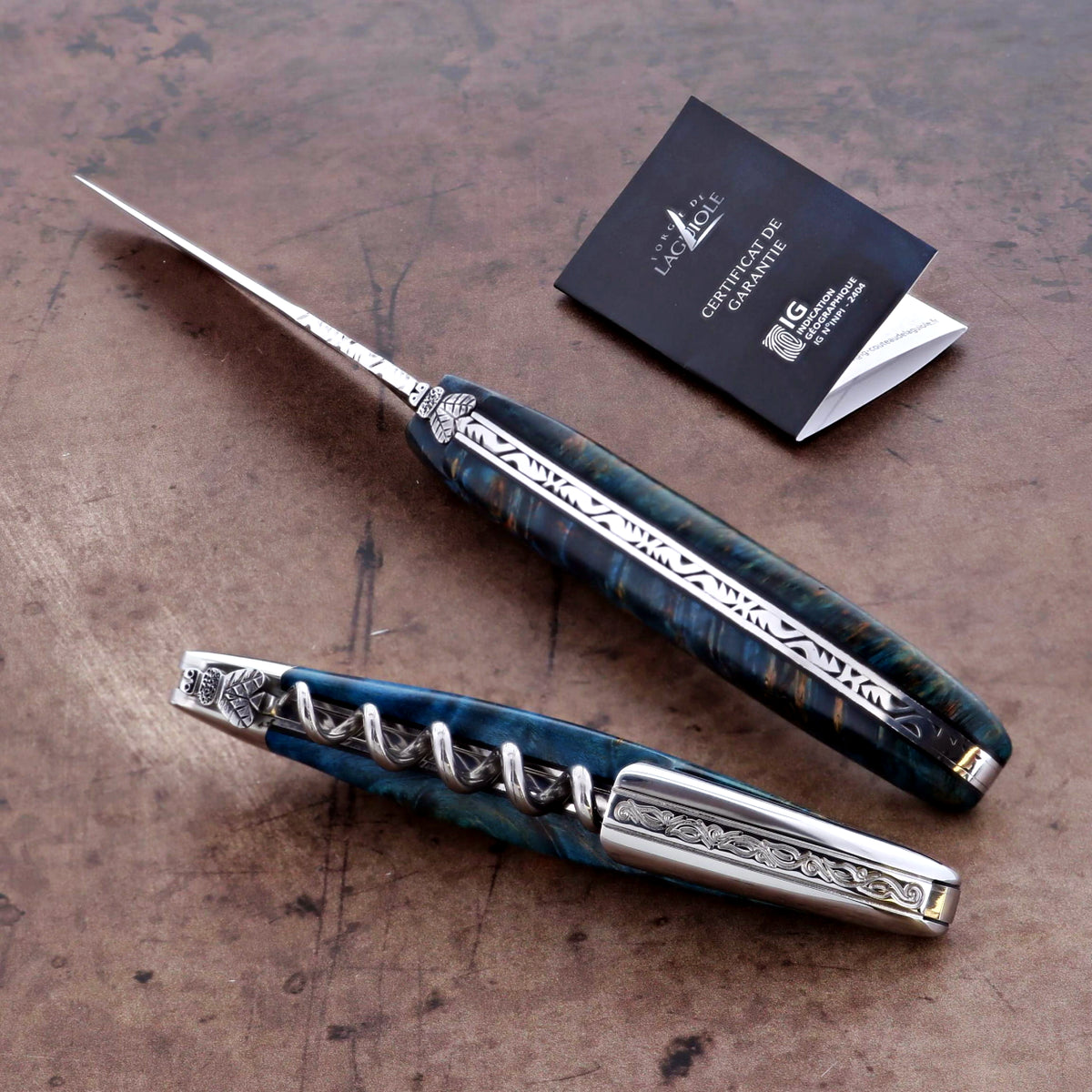Two ornate laguiole with decorative handles on a textured surface, accompanied by a branded card.
