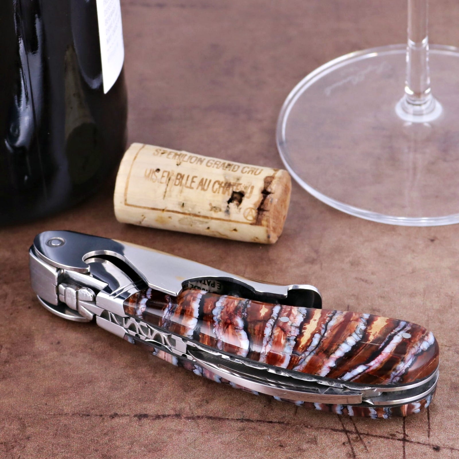 Laguiole Magnum Guilloche Mammoth Tooth corkscrew, close-up of molten earth handle, wine bottle, cork, glass.