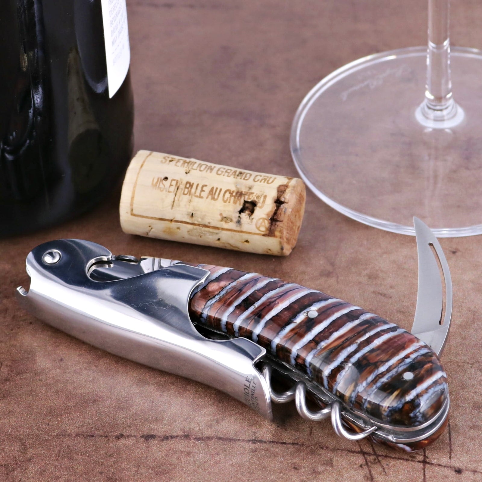 Laguiole Magnum Guilloche Mammoth Tooth corkscrew, close-up of molten earth handle, wine bottle, cork, glass.