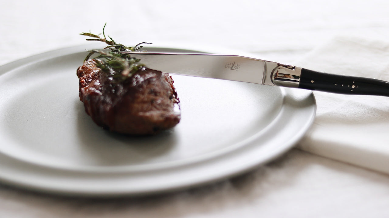 forge de laguiole black horn steak knife on a white plate next to a filet mignon steak