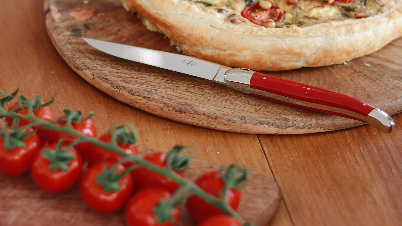 https://www.laguiole-imports.com/cdn/shop/files/forge_de_laguiole_steak_knife_with_red_handle_next_to_tomato_and_pizza_1600x.jpg?v=1699543904